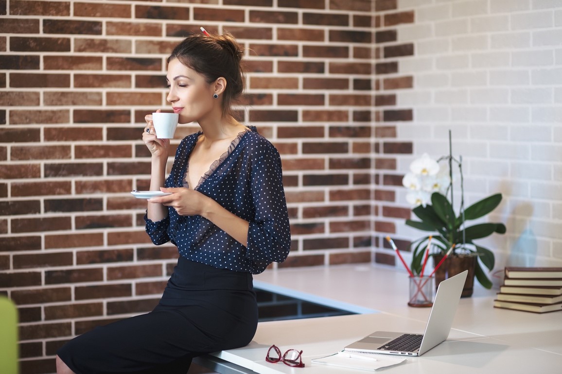 Une femme à son bureau, en jupe crayon, en train de boire un café