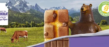 Un ours tient une madeleine chocolatée Milka devant un fond Alpin