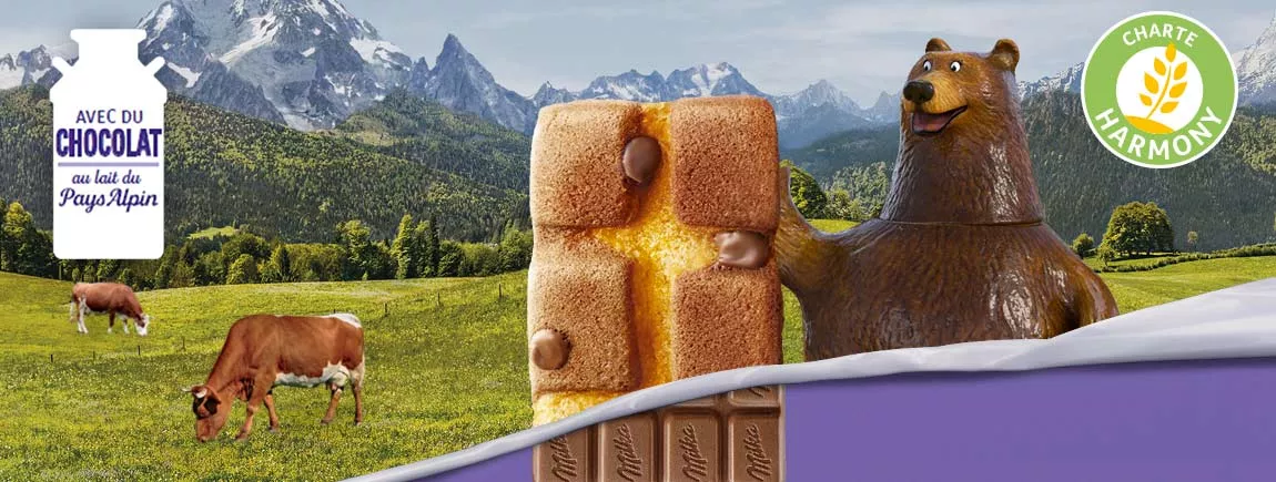 Un ours tient une madeleine chocolatée Milka devant un fond Alpin